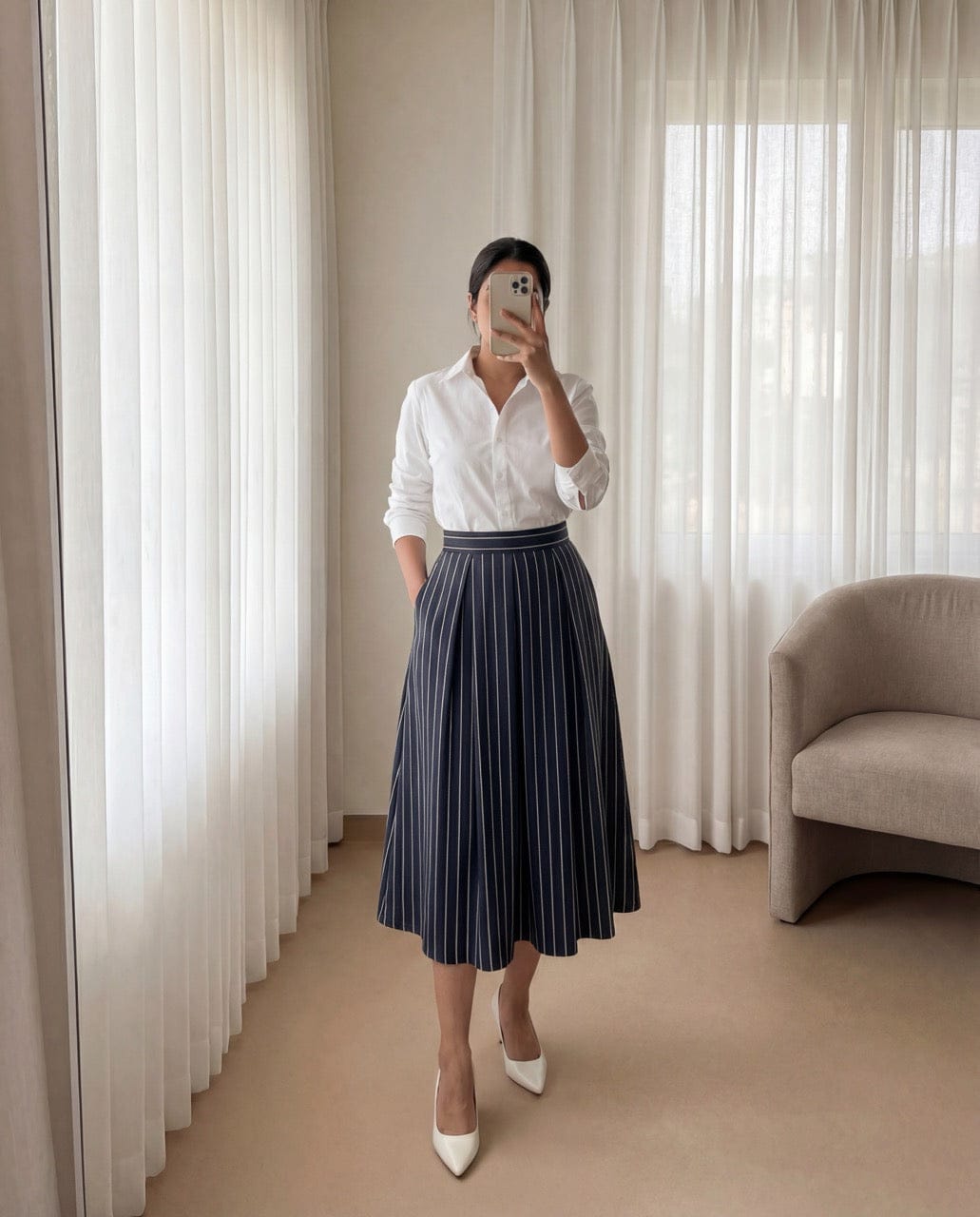 Person wearing a white shirt and navy pleated skirt in a room with beige walls and a chair.